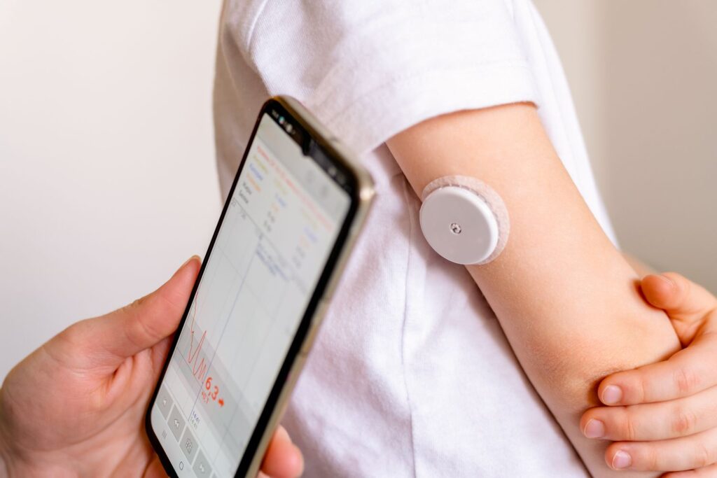 A child wearing a glucose sensor on their arm while a smartphone app displays real-time blood sugar levels and trends.