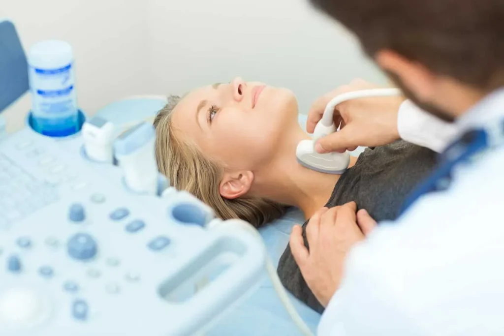 A doctor performing a thyroid ultrasound on a female patient using a transducer probe on her neck in a clinical setting.