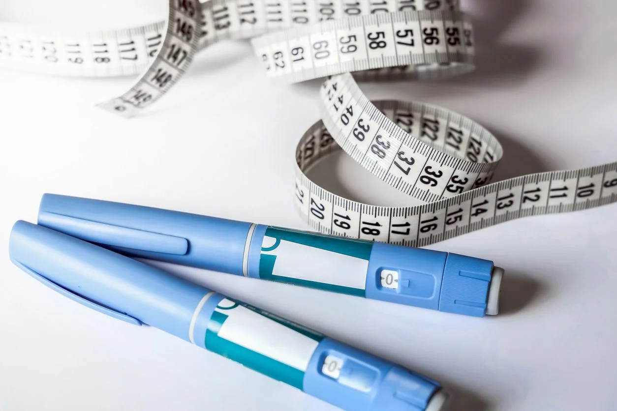 Two blue medical injection pens lying on a white surface next to a coiled white measuring tape.